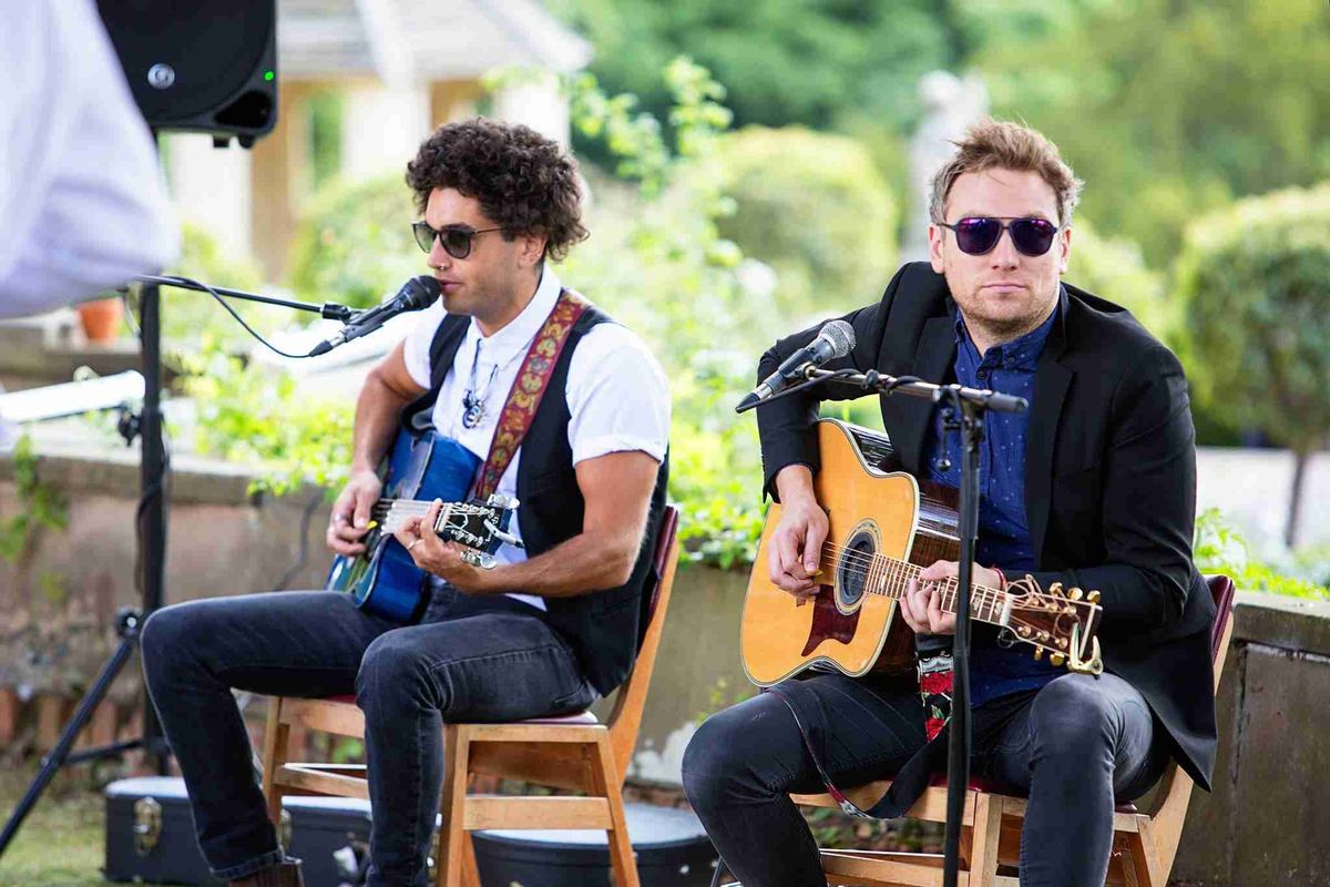 Two members of the band playing acoustic guitars in a garden